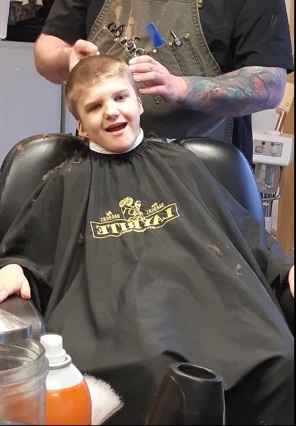 The student is wearing a black cape and grinning as he gets a haircut from a barber who is using trimmers.