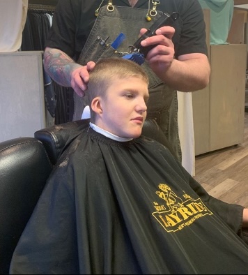 A barber uses trimmers to cut the top of the student's hair. 