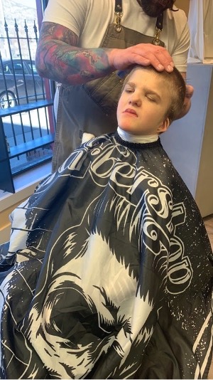 The student leans back while a barber uses trimmers to cut the hair on the back of the student's head. 