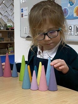 Student wearing glasses and arranging conical plastic shapes on her desk. She is touching the point of the purple cone. 
