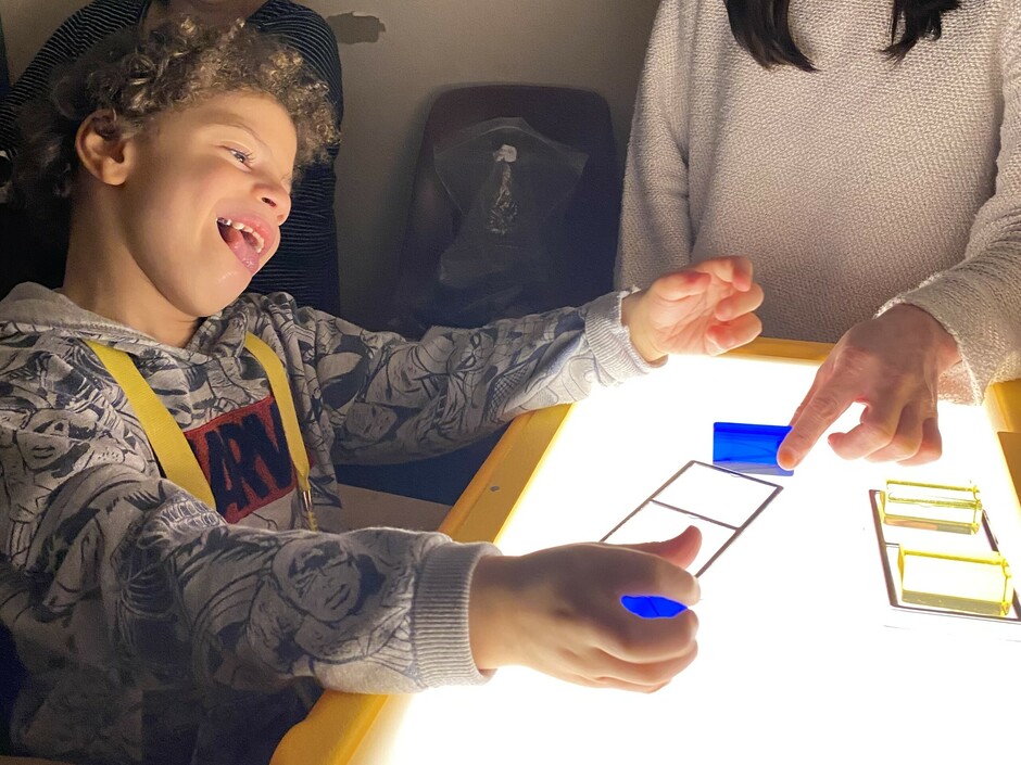 A student laughs while sitting in front of a light box. The student has a translucent blue block in his hand. His Intervenor has a matching block.