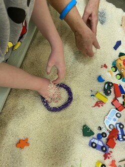 Intervenor and student using their hands to play with rice. Small, colourful vehicles and dinosaurs are available, as well as a container for scooping.