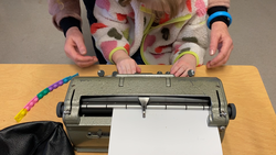 Student pressing keys on a Perkins braille writer independently. Her Intervenor's hands are nearby in case she needs support.