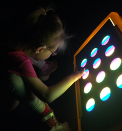 Student sitting on the floor looking multi coloured circles and black background. She is independently touching the light box with her right hand. 